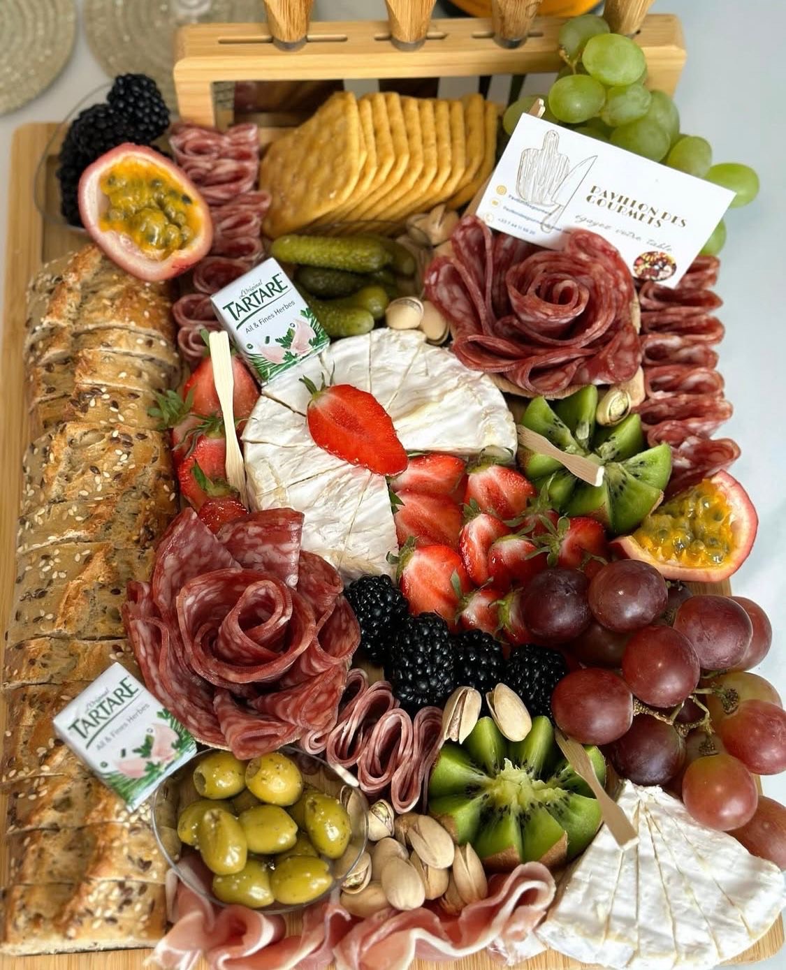 Plateau de charcuterie composé de viandes variées, fromages, fruits frais, noix, olives, crackers et pain, soigneusement disposés sur un plateau en bois avec une carte de visite portant l'inscription Pavillon des Gourmets, dans un environnement lumineux et accueillant